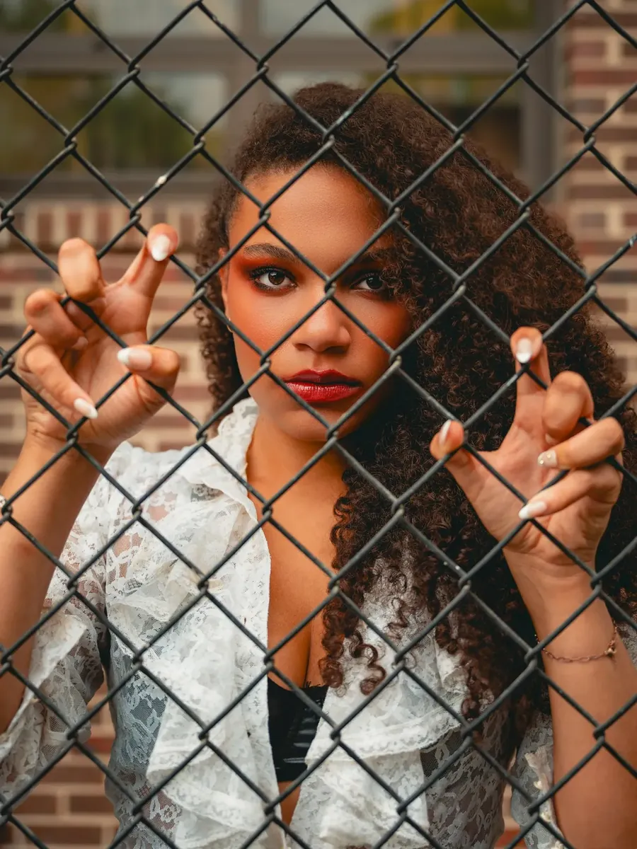 Tig stares out through a chain-link fence, hands gripping the wire.