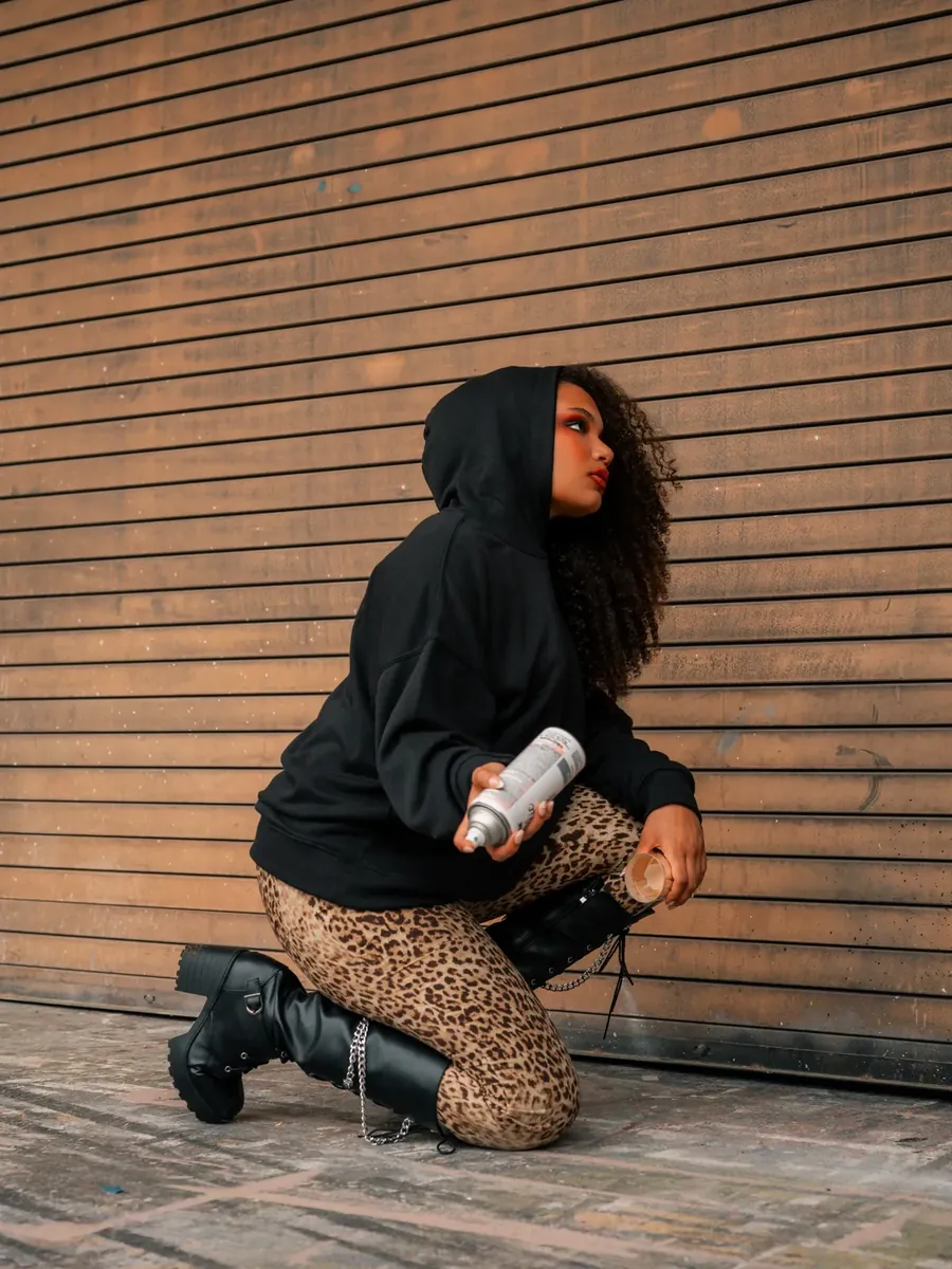 A hooded figure kneels with a spray-paint can against a shuttered storefront.